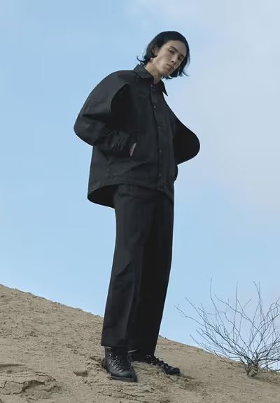 A man standing on a sandy hill wearing a black Konundrum jacket with matching black pants and boots