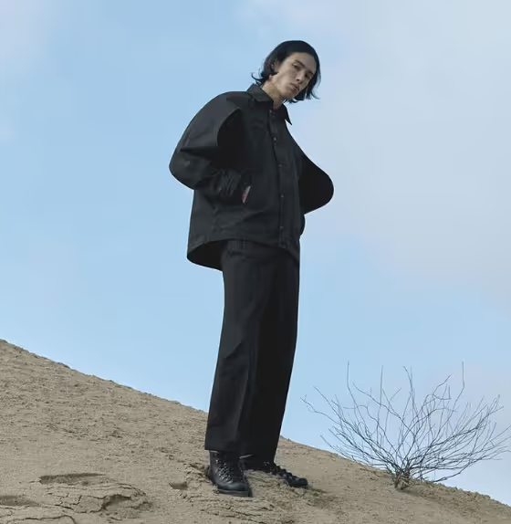 A man standing on a sandy hill wearing a black Konundrum jacket with matching black pants and boots