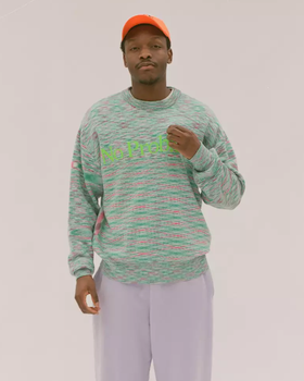 A man in a colorful Aries green sweater, lavender pants and an orange hat