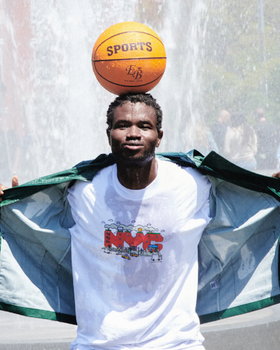A man standing in a sprinkler with a basketball balanced on his head wearing Only NY t-shirt and jacket
