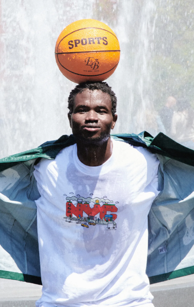 A man standing in a sprinkler with a basketball balanced on his head wearing Only NY t-shirt and jacket