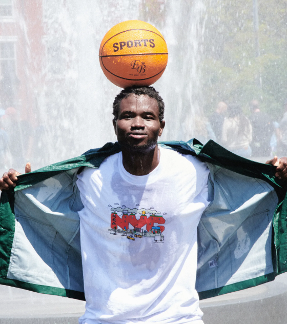A man standing in a sprinkler with a basketball balanced on his head wearing Only NY t-shirt and jacket