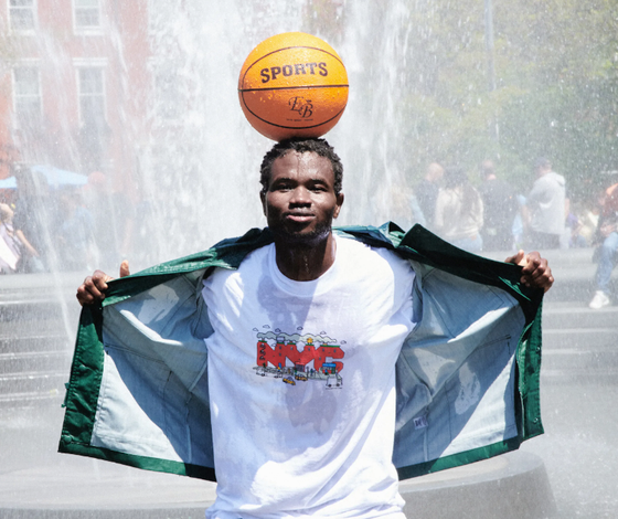 A man standing in a sprinkler with a basketball balanced on his head wearing Only NY t-shirt and jacket