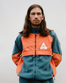 A man with long brown hair wearing an orange and green 90s style Palace fleece jacket