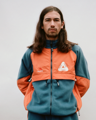 A man with long brown hair wearing an orange and green 90s style Palace fleece jacket
