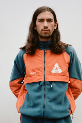A man with long brown hair wearing an orange and green 90s style Palace fleece jacket
