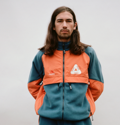 A man with long brown hair wearing an orange and green 90s style Palace fleece jacket