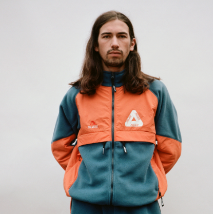 A man with long brown hair wearing an orange and green 90s style Palace fleece jacket
