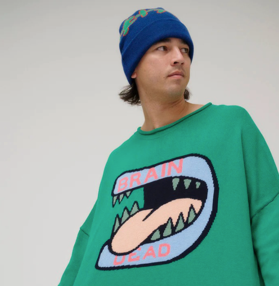 A man wearing a green Brain Dead Sweater with a drawing of a monsters mouth with it's tongue out on it and a blue beanie