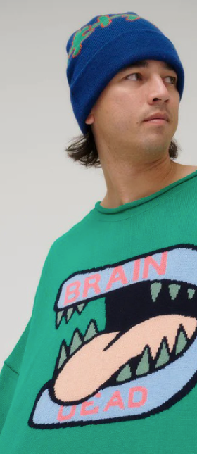 A man wearing a green Brain Dead Sweater with a drawing of a monsters mouth with it's tongue out on it and a blue beanie