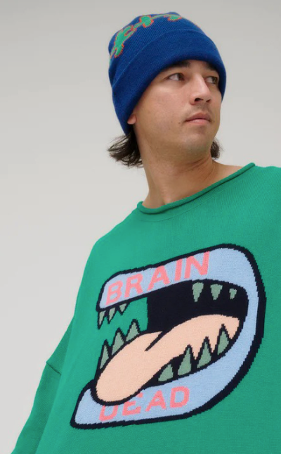 A man wearing a green Brain Dead Sweater with a drawing of a monsters mouth with it's tongue out on it and a blue beanie