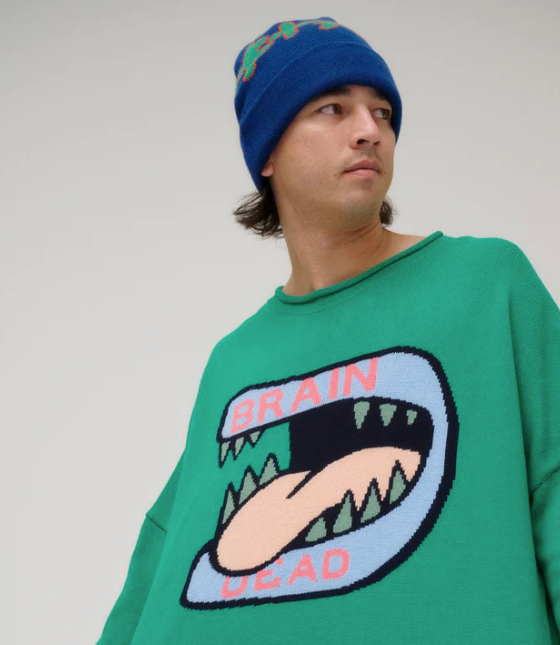 A man wearing a green Brain Dead Sweater with a drawing of a monsters mouth with it's tongue out on it and a blue beanie