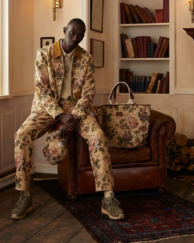 A man sitting on a desk wearing a beige Kith floral printed jacket and matching pants and bag
