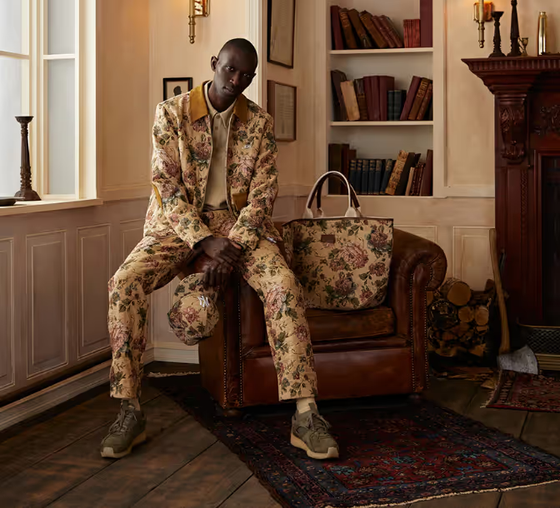 A man sitting on a desk wearing a beige Kith floral printed jacket and matching pants and bag