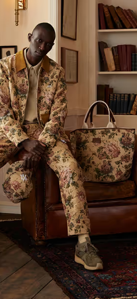 A man sitting on a desk wearing a beige Kith floral printed jacket and matching pants and bag