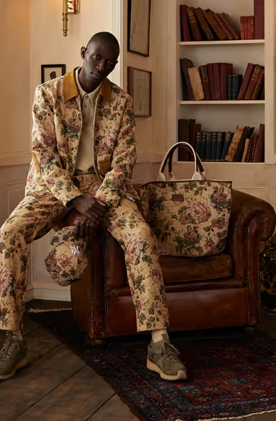 A man sitting on a desk wearing a beige Kith floral printed jacket and matching pants and bag