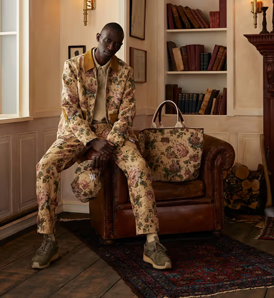 A man sitting on a desk wearing a beige Kith floral printed jacket and matching pants and bag