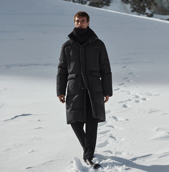 A man bundled up wearing a dark grey COS jacket, scarf and pants walking in the snow