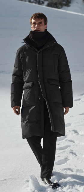 A man bundled up wearing a dark grey COS jacket, scarf and pants walking in the snow