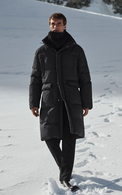 A man bundled up wearing a dark grey COS jacket, scarf and pants walking in the snow