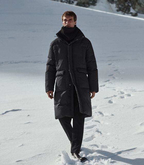 A man bundled up wearing a dark grey COS jacket, scarf and pants walking in the snow