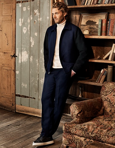 A man with blonde curly hair in a lodge cabin waring a navy blue Club Monaco jacket and pants, with a beige turtleneck