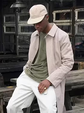 A man wearing an Asos biege jacket, green t-shirt, white pants and a matching white hat