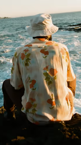 A man sitting while looking out into the ocean wearing a floral General Admission button up and a white bucket hat