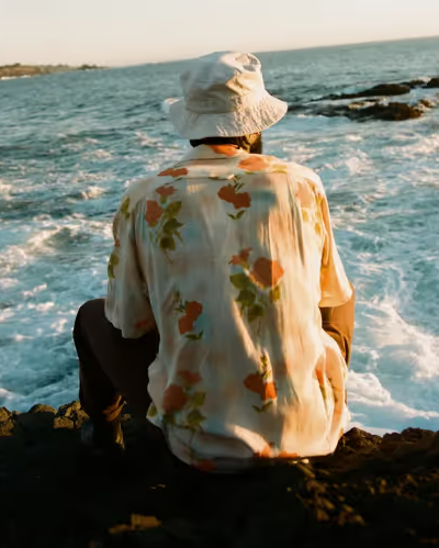 A man sitting while looking out into the ocean wearing a floral General Admission button up and a white bucket hat