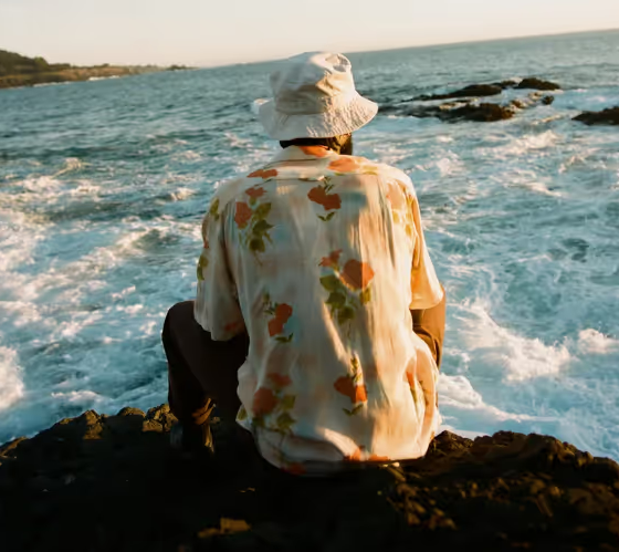A man sitting while looking out into the ocean wearing a floral General Admission button up and a white bucket hat