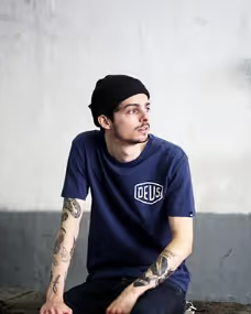 A man looking off into the distance wearing a Navy Blue shirt with a large Deus logo and matching beanie