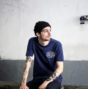A man looking off into the distance wearing a Navy Blue shirt with a large Deus logo and matching beanie