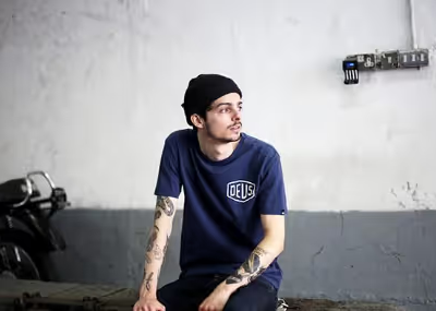A man looking off into the distance wearing a Navy Blue shirt with a large Deus logo and matching beanie