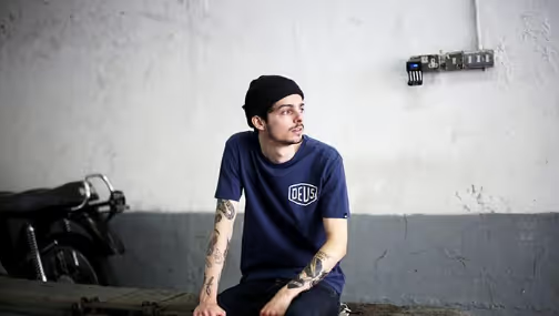 A man looking off into the distance wearing a Navy Blue shirt with a large Deus logo and matching beanie