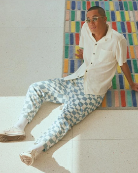 A man sitting on the floor wearing light blue and white Banks Journal checkered pants, a beige button up and sunglasses