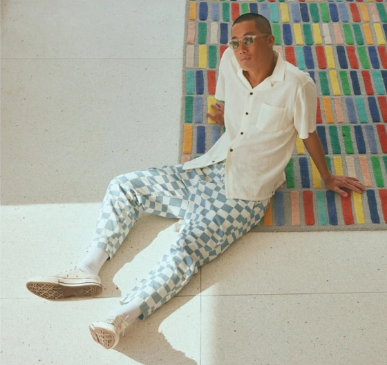 A man sitting on the floor wearing light blue and white Banks Journal checkered pants, a beige button up and sunglasses