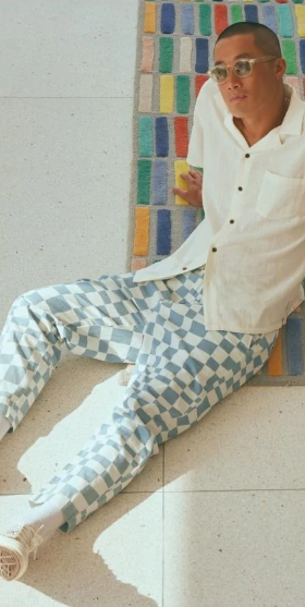 A man sitting on the floor wearing light blue and white Banks Journal checkered pants, a beige button up and sunglasses