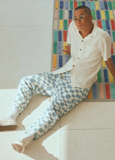 A man sitting on the floor wearing light blue and white Banks Journal checkered pants, a beige button up and sunglasses