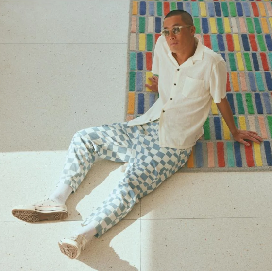 A man sitting on the floor wearing light blue and white Banks Journal checkered pants, a beige button up and sunglasses