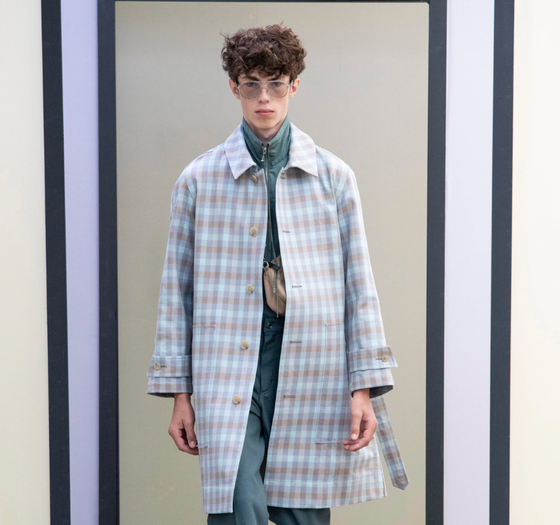 A man wearing a baby blue checkered print Maison Kitsune trench coat and clear vintage glasses