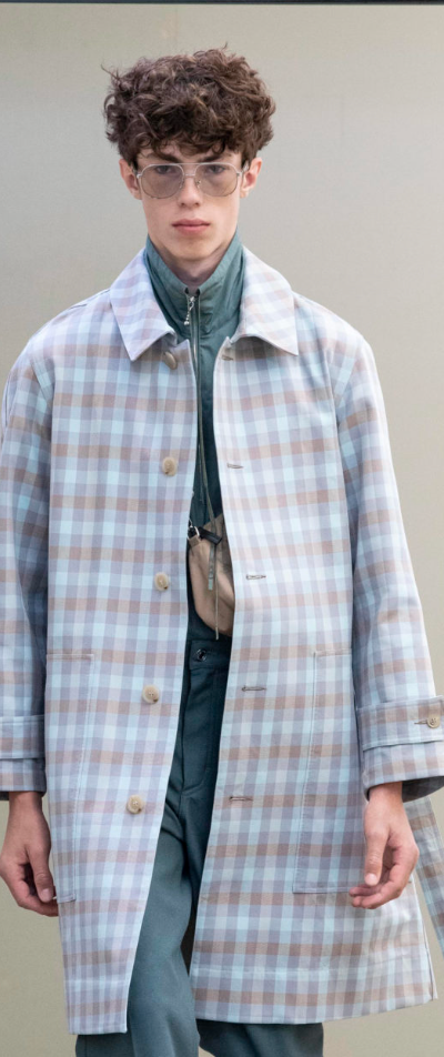 A man wearing a baby blue checkered print Maison Kitsune trench coat and clear vintage glasses