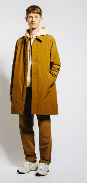 A man wearing a long light brown trench coat with matching pants and Oatmeal Sweatshirt along with New Balance Dad Shoes