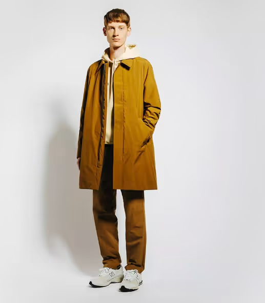A man wearing a long light brown trench coat with matching pants and Oatmeal Sweatshirt along with New Balance Dad Shoes
