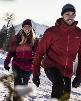 A couple hiking in the snow wearing matching Maroon Red Vaude Jackets, black pants and a matching beanies