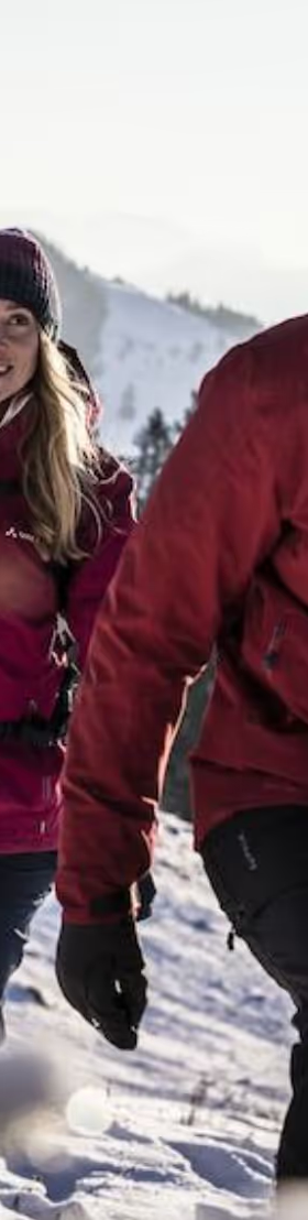 A couple hiking in the snow wearing matching Maroon Red Vaude Jackets, black pants and a matching beanies