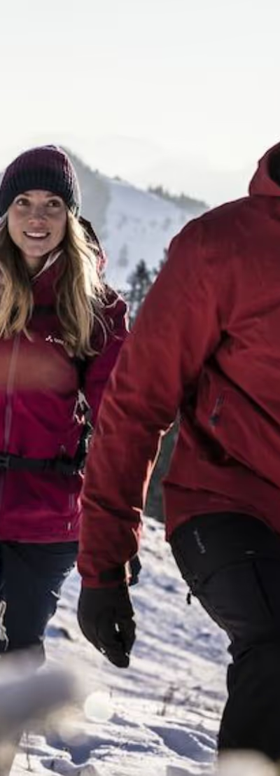 A couple hiking in the snow wearing matching Maroon Red Vaude Jackets, black pants and a matching beanies