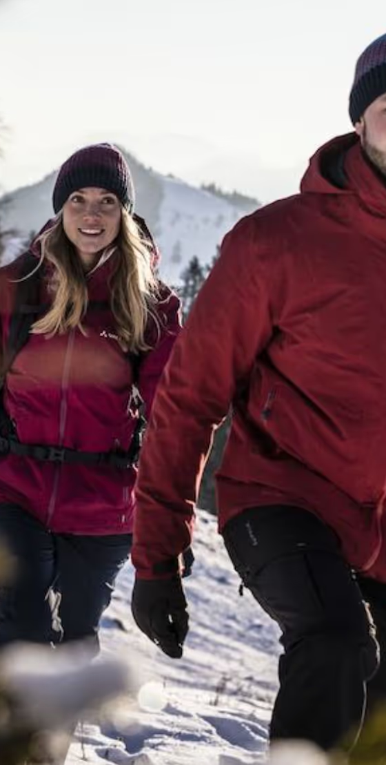 A couple hiking in the snow wearing matching Maroon Red Vaude Jackets, black pants and a matching beanies