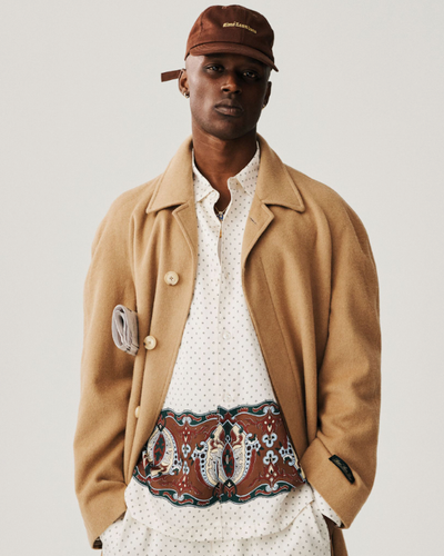 A man wearing a brown Aime Leon Dore trench coat and white button up with a dark brown hat