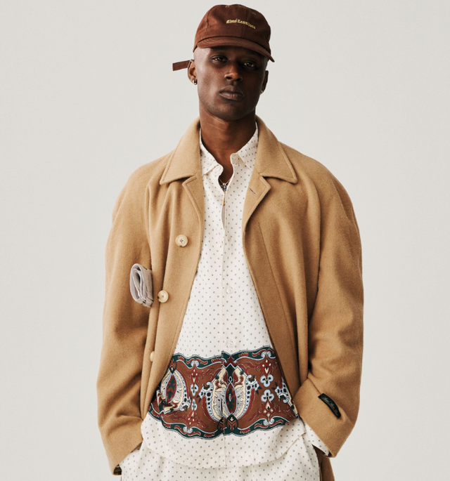 A man wearing a brown Aime Leon Dore trench coat and white button up with a dark brown hat