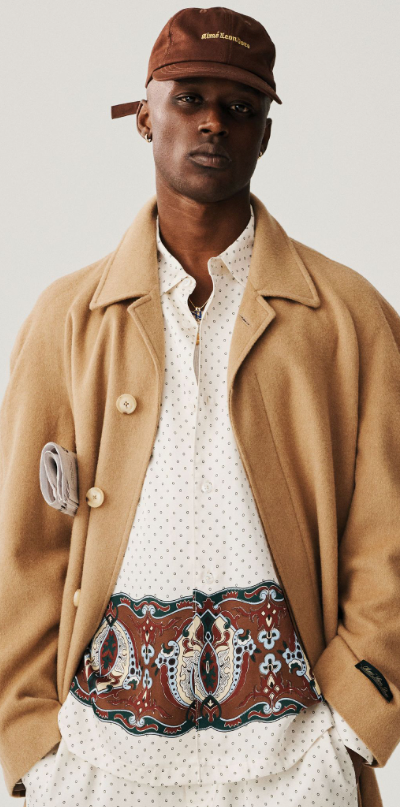 A man wearing a brown Aime Leon Dore trench coat and white button up with a dark brown hat